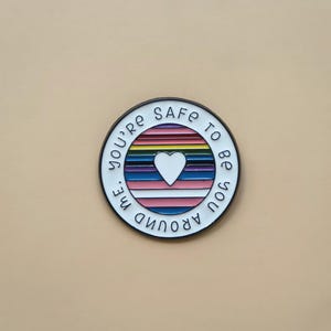 Puede incluir: Un pin redondo de esmalte con borde negro. El pin presenta un diseño de rayas de arcoíris con un corazón blanco en el centro. El texto "You're safe to be you around me" está escrito alrededor del borde del pin.