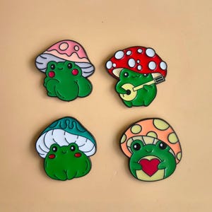 Op de afbeelding: Vier grillige emaille pins met groene kikkers onder kleurrijke paddenstoelenkappen. Eén kikker speelt gitaar, een ander houdt een hart vast. De pins hebben zwarte contouren en staan tegen een neutrale achtergrond.