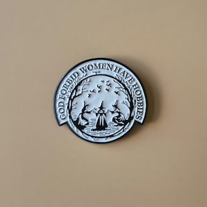 May include: A black and white circular pin with the text "GOD FORBID WOMEN HAVE HOBBIES". The pin features a graphic of three witches in a circle, with trees and birds. The pin is set against a tan background.
