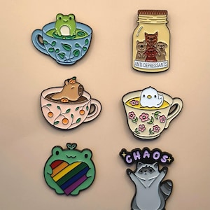 May include: Six enamel pins featuring a green frog in a blue teacup, a jar of dogs labeled ANTI DEPRESSANTS, a capybara in a floral teacup, a white bird in a floral teacup, a green frog holding a rainbow heart, and a raccoon with the word CHAOS.