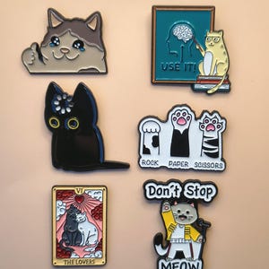 May include: A collection of six enamel pins featuring cat-themed designs: a crying cat giving a thumbs up, a cat pointing to a brain with the text USE IT, a black cat, a cat paw rock paper scissors graphic, a Lovers tarot card, and a cat singing.