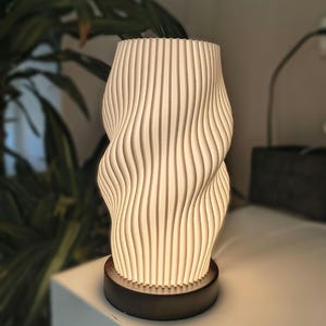Op de afbeelding: Een cilindrische tafellamp met een witte, verticaal geribbelde kap met een golvende textuur. De lamp wordt van binnenuit verlicht, verspreidt een zachte gloed en is gemonteerd op een ronde, donkerbruine houten voet.