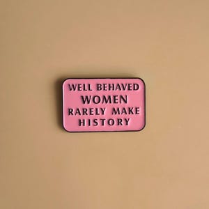Op de afbeelding: Een rechthoekige emaille speld met een roze achtergrond en een zwarte rand. De speld bevat de zwarte tekst "WELL BEHAVED WOMEN RARELY MAKE HISTORY" gecentreerd in vier regels. Het ontwerp heeft afgeronde hoeken en een gladde, gepolijste afwerking.
