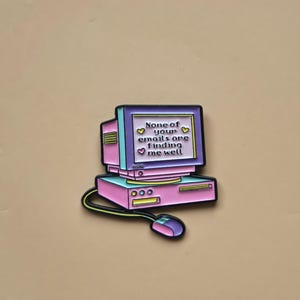 Op de afbeelding: Een retro computer pin in roze en paars. Het scherm toont de tekst "None of your emails are finding me well" met hartjes. De pin heeft een muis en een snoer. Een nostalgisch ontwerp.