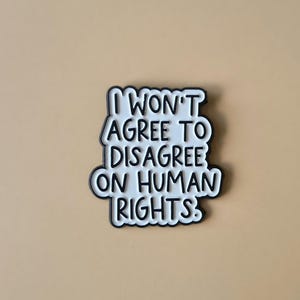 Op de afbeelding: Een wit en zwart speldje met de tekst "I WON'T AGREE TO DISAGREE ON HUMAN RIGHTS." De speld heeft een zwarte rand en staat tegen een beige achtergrond.