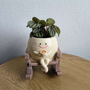 Könnte beinhalten: Ein skurriler, cremefarbener Keramik-Pflanztopf in Form eines lächelnden Charakters, der in einem Holzschaukelstuhl sitzt. Der Topf enthält eine kleine grüne Pflanze mit gemusterten Blättern. Die Figur hält eine kleine Tasse.
