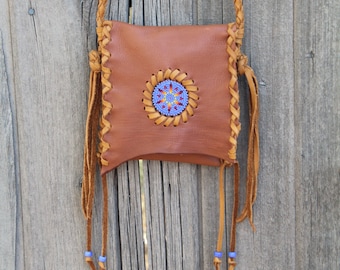May include: A small brown leather bag with a beaded design and fringe. The bag is hanging on a wooden fence.