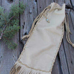 May include: A beige leather drawstring pouch featuring braided side seams and long fringe at the bottom. The bag includes a braided leather cord closure with a small light blue bead accent near the top opening, providing a secure and functional design.