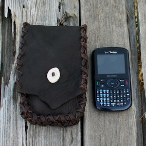 Op de afbeelding: Een bruine leren tas met een gevlochten rand en een bottenknop. De tas ligt naast een zwarte mobiele telefoon met een QWERTY-toetsenbord en de tekst "Verizon" en "Pantech" erop.