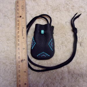 K&ouml;nnte beinhalten: Ein kleiner schwarzer Lederbeutel mit einem Design aus t&uuml;rkisfarbenen und roten Perlen. Der Beutel ist an einem schwarzen Lederband befestigt.