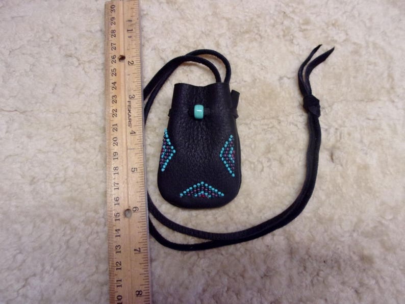 K&ouml;nnte beinhalten: Ein kleiner schwarzer Lederbeutel mit einem Design aus t&uuml;rkisfarbenen und roten Perlen. Der Beutel ist an einem schwarzen Lederband befestigt.