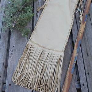 May include: A beige leather pouch featuring braided side lacing, a drawstring closure with light blue beads, and long fringe at the bottom. A wooden yardstick is placed alongside the pouch, showing a length of approximately 18 inches.