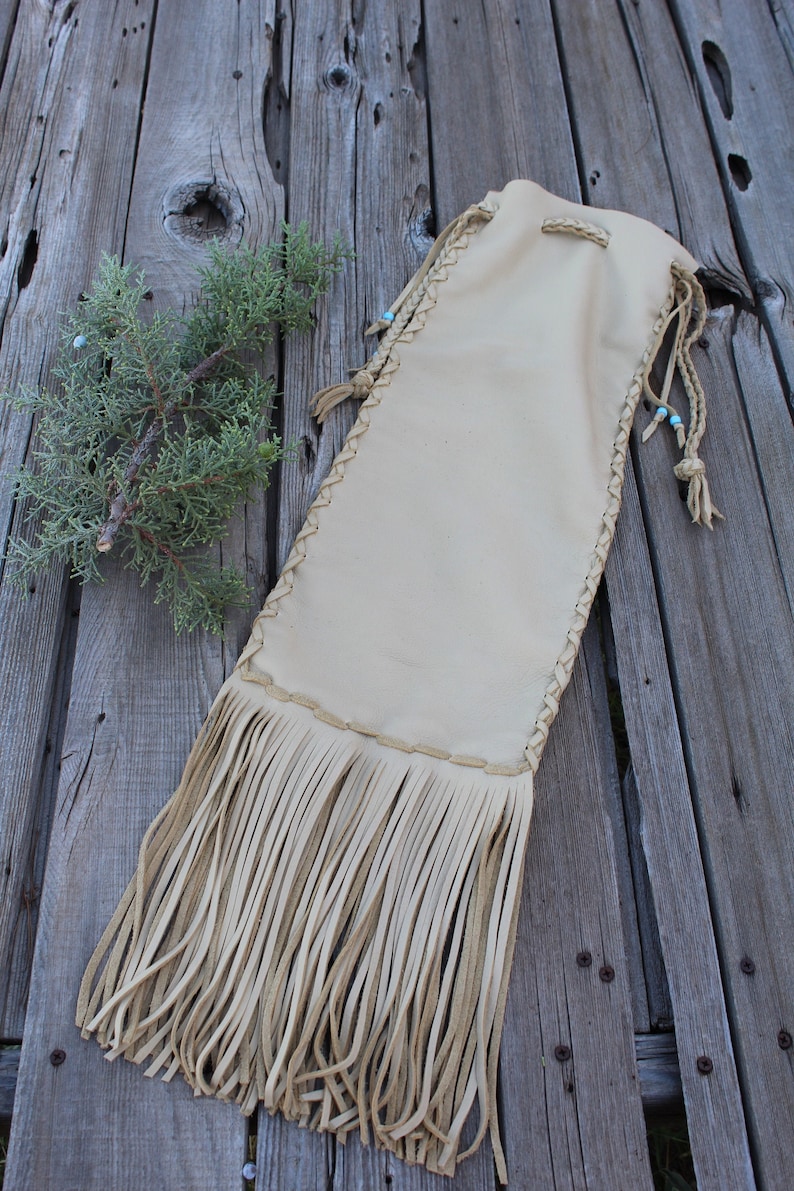 May include: A beige leather pouch featuring long fringe at the bottom, braided side detailing, and drawstring closures with small light blue beads. The pouch has a smooth texture and is constructed with visible stitching along the edges.