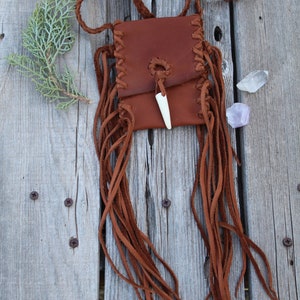 Pu&ograve; includere: Una piccola borsa in pelle marrone con un bordo a frangia e un ciondolo in osso. La borsa &egrave; appesa a una tracolla in pelle intrecciata.