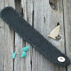 May include: A black leather bracelet with a stitched design and a silver button. The bracelet is lying on a weathered wooden surface with three turquoise stones.