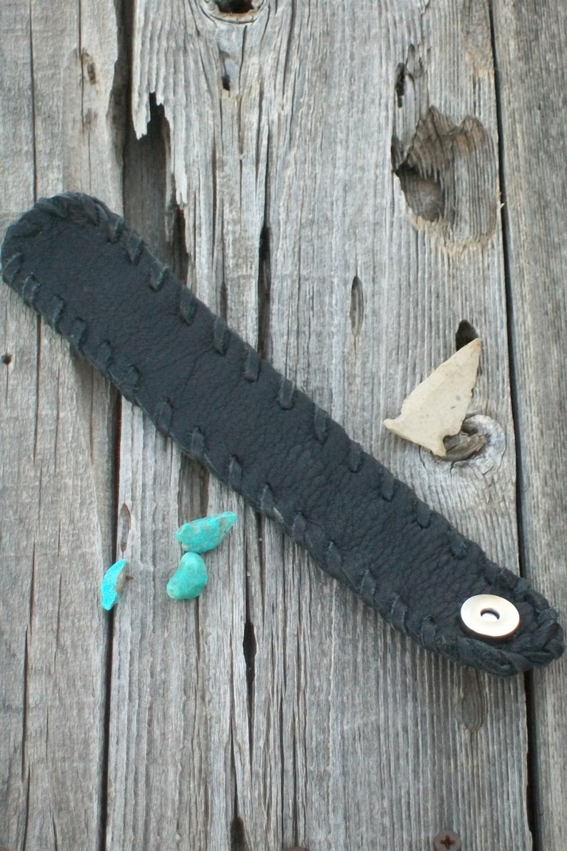 May include: A black leather bracelet with a stitched design and a silver button. The bracelet is lying on a weathered wooden surface with three turquoise stones.