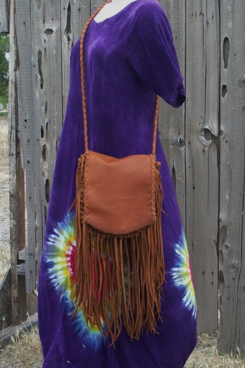 May include: A brown leather crossbody bag with long fringe. The bag has a braided strap and is worn over a purple tie-dye dress. The bag's leather is a warm, natural tone, and the fringe hangs down.
