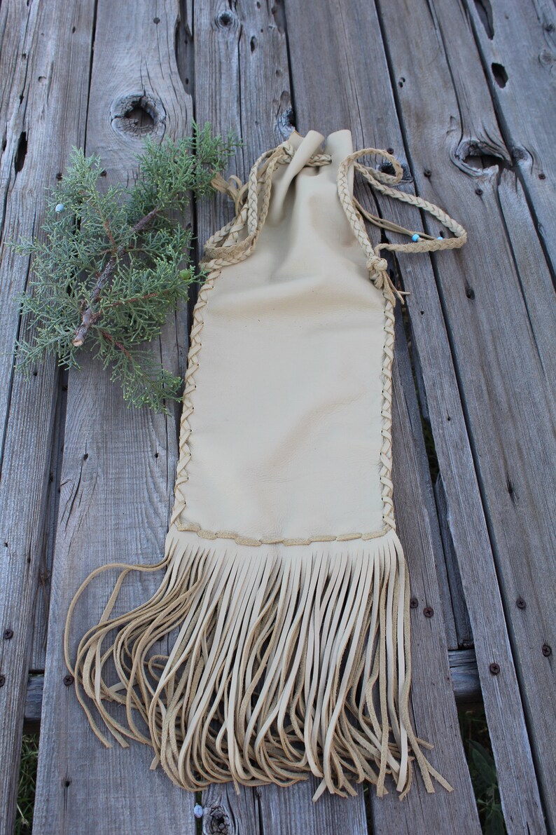 May include: A beige leather drawstring pouch featuring braided leather side seams and a long fringe trim at the bottom. The bag includes braided leather straps for closure and is accented with small blue beads on the ends of the drawstrings.