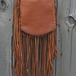 May include: A brown leather crossbody bag with long fringe. The bag has a flap closure and a braided strap. The fringe hangs from the bottom of the bag, adding a bohemian flair. The bag is hanging against a weathered wooden background.