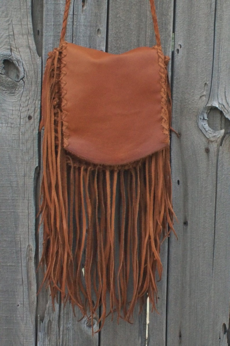 May include: A brown leather crossbody bag with long fringe. The bag has a flap closure and a braided strap. The fringe hangs from the bottom of the bag, adding a bohemian flair. The bag is hanging against a weathered wooden background.