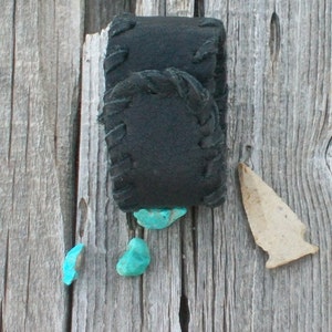 May include: A black leather pouch with a decorative stitch pattern, attached to a wooden surface. Four turquoise stones are scattered around the pouch.