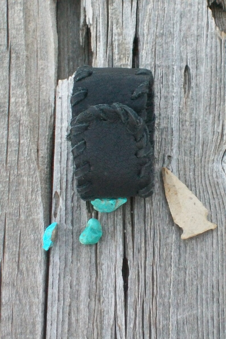 May include: A black leather pouch with a decorative stitch pattern, attached to a wooden surface. Four turquoise stones are scattered around the pouch.