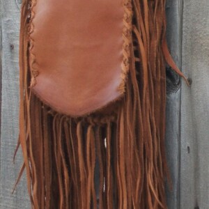May include: A brown leather shoulder bag with long, flowing fringe. The bag has a rectangular body with a rounded bottom and is adorned with decorative stitching along the edges. The fringe hangs down the length of the bag.