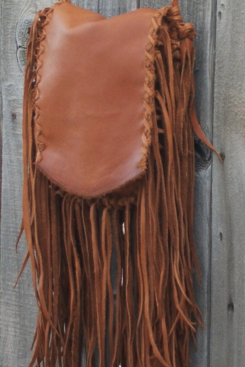 May include: A brown leather shoulder bag with long, flowing fringe. The bag has a rectangular body with a rounded bottom and is adorned with decorative stitching along the edges. The fringe hangs down the length of the bag.