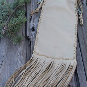 May include: A beige leather drawstring pouch featuring braided side seams, long fringe at the bottom, and thin leather cords with small light blue beads at the top. The pouch has a smooth, matte finish and a rectangular shape with rounded corners.