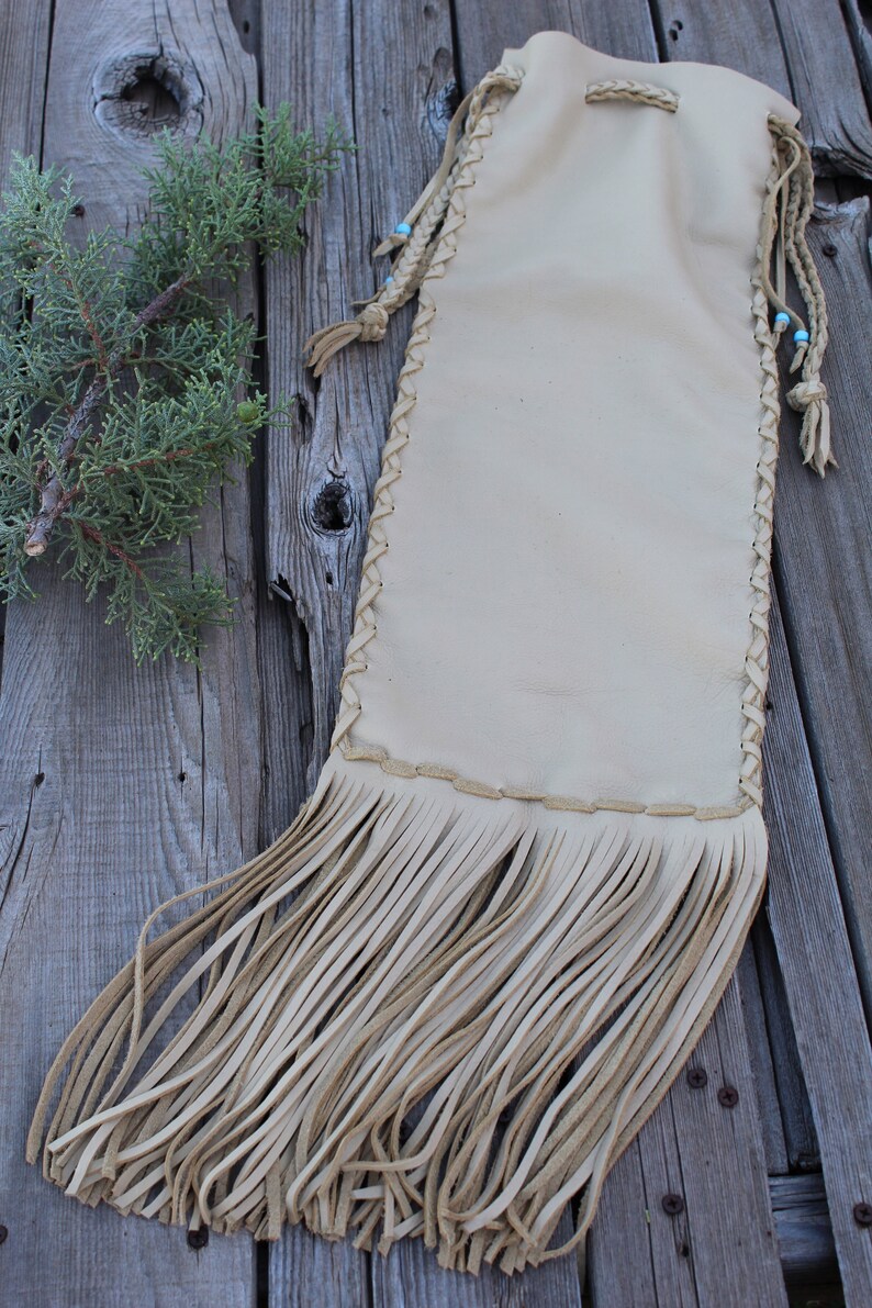 May include: A beige leather drawstring pouch featuring braided side seams, long fringe at the bottom, and thin leather cords with small light blue beads at the top. The pouch has a smooth, matte finish and a rectangular shape with rounded corners.