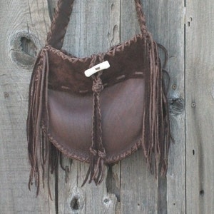 May include: A brown leather shoulder bag with fringe detailing and a bone button closure. The bag is hanging on a weathered wooden surface.