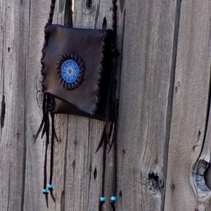 May include: A brown leather purse with a beaded design and fringe. The purse is hanging on a wooden fence.