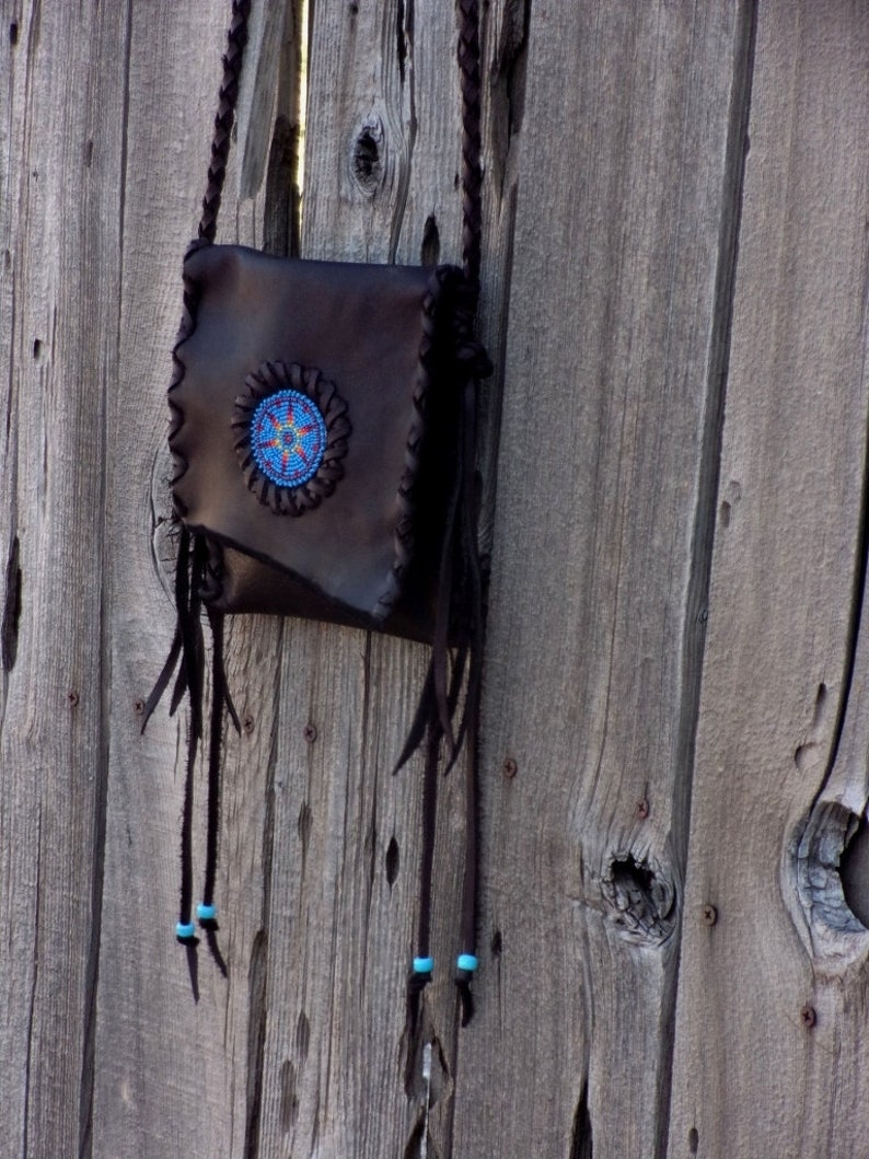 May include: A brown leather purse with a beaded design and fringe. The purse is hanging on a wooden fence.
