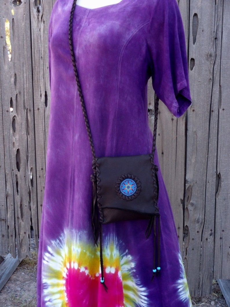 May include: A brown leather crossbody bag with a beaded design and fringe accents. The bag is worn over a purple tie-dye dress.