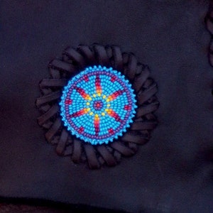 May include: A close-up of a blue, red, yellow, and white beaded design on a black leather background. The design is circular and has a starburst pattern.