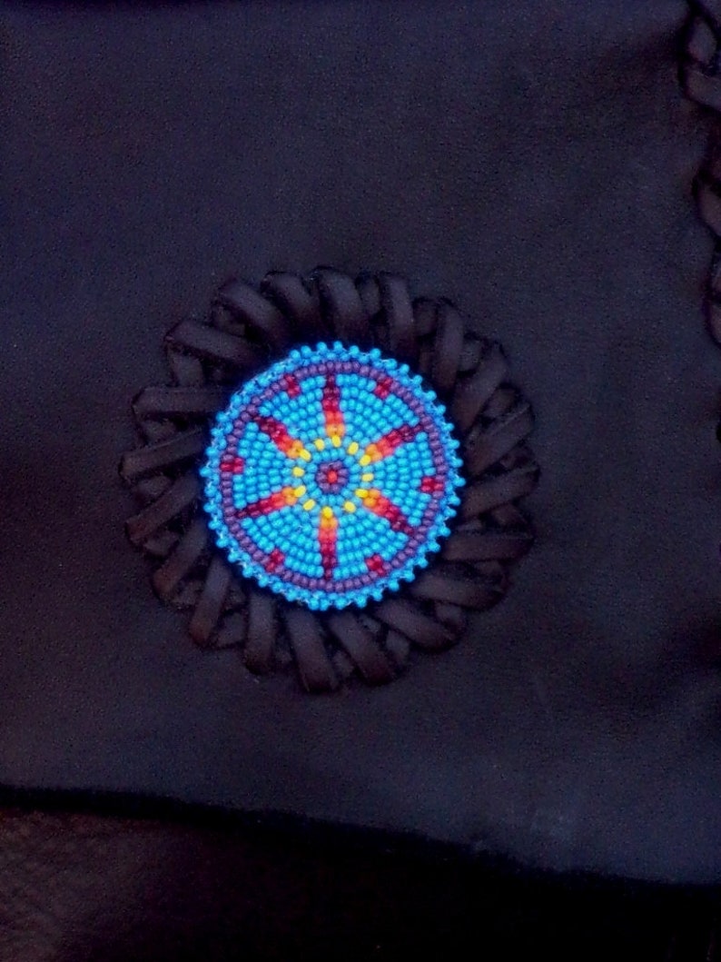May include: A close-up of a blue, red, yellow, and white beaded design on a black leather background. The design is circular and has a starburst pattern.