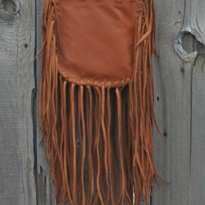 May include: A brown leather crossbody bag with long fringe along the bottom. The bag has a braided strap and is against a weathered wooden background. The bag's design is simple, with a rectangular body and a gathered top.