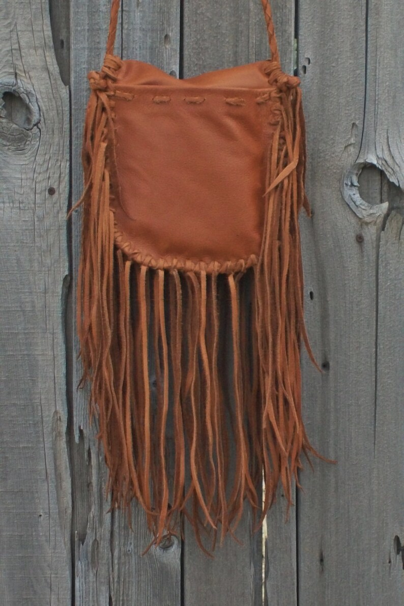 May include: A brown leather crossbody bag with long fringe along the bottom. The bag has a braided strap and is against a weathered wooden background. The bag's design is simple, with a rectangular body and a gathered top.
