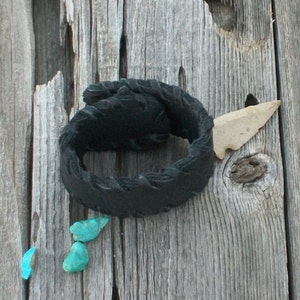 May include: A black leather bracelet with a braided design, a small arrowhead, and three turquoise stones on a weathered wooden surface.