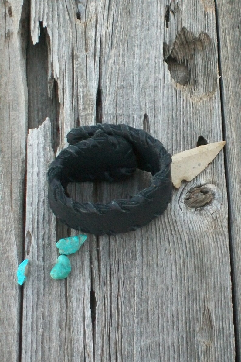 May include: A black leather bracelet with a braided design, a small arrowhead, and three turquoise stones on a weathered wooden surface.