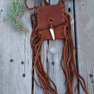Pu&ograve; includere: Una piccola borsa in pelle marrone con bordi a frangia e chiusura in osso. La borsa &egrave; appesa a una cinghia in pelle intrecciata e poggia su una superficie in legno.