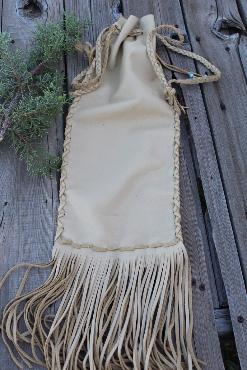 May include: A cream-colored leather drawstring pouch featuring braided leather straps and a long fringe detail along the bottom edge. The pouch is constructed with visible braided stitching along the sides and includes a small blue bead accent.