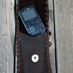 Op de afbeelding: Een bruine leren telefoonhoes met een gevlochten ontwerp en een drukknoop sluiting. Een zwarte telefoon met een QWERTY-toetsenbord zit in de hoes.
