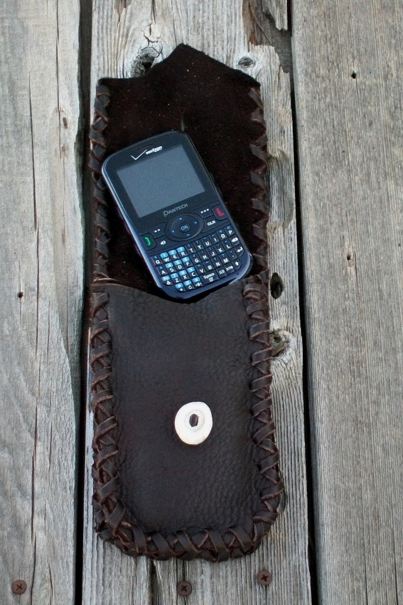 Op de afbeelding: Een bruine leren telefoonhoes met een gevlochten ontwerp en een drukknoop sluiting. Een zwarte telefoon met een QWERTY-toetsenbord zit in de hoes.