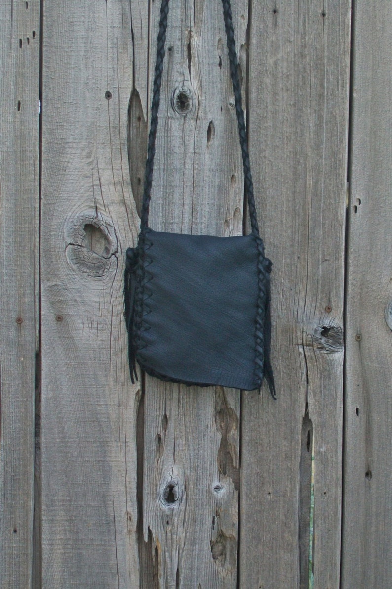 K&ouml;nnte beinhalten: Eine schwarze Leder-Umh&auml;ngetasche mit geflochtenem Riemen und Fransen. Die Tasche h&auml;ngt an einem Holzzaun.