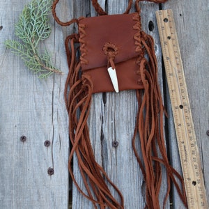 Pu&ograve; includere: Una borsa in pelle marrone con frange e un ciondolo in osso. La borsa &egrave; appesa a una tracolla in pelle intrecciata.