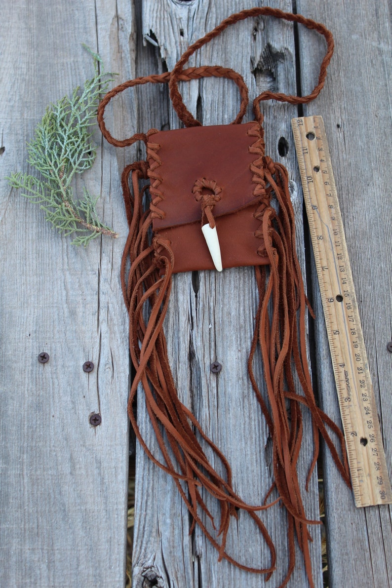 Pu&ograve; includere: Una borsa in pelle marrone con frange e un ciondolo in osso. La borsa &egrave; appesa a una tracolla in pelle intrecciata.