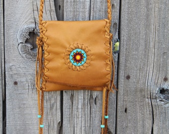 May include: A brown leather crossbody bag with a beaded sun design and turquoise accents. The bag has a long strap and fringe details.