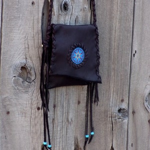 May include: A brown leather bag with a beaded design and turquoise accents. The bag is hanging on a wooden wall.
