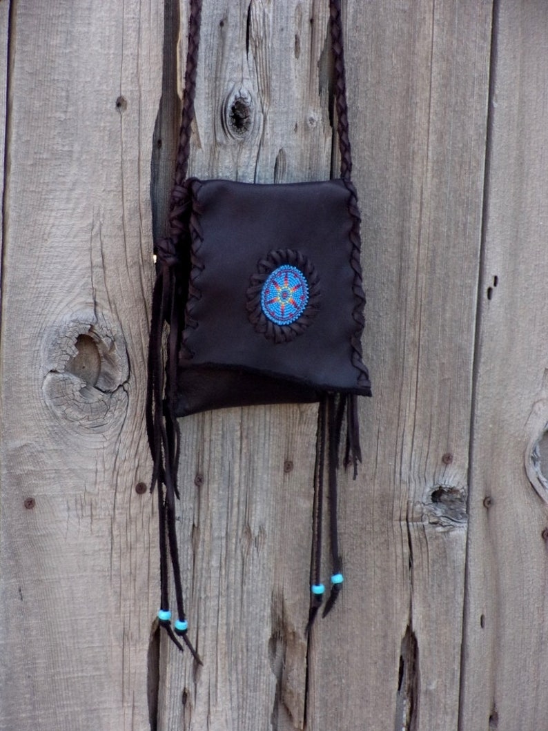 May include: A brown leather bag with a beaded design and turquoise accents. The bag is hanging on a wooden wall.